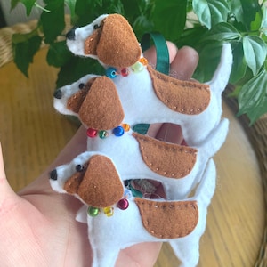 May include: Three handmade felt dog ornaments in white with brown patches on the ears and back. Each dog features a small black bead eye and a collar made of colourful, translucent plastic beads. The ornaments are attached to a dark green ribbon loop.