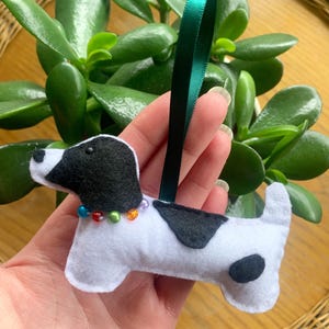 May include: A handmade felt dog ornament with a white body, black spots, and a black head. It has a colorful beaded collar and a green ribbon for hanging. The ornament is approximately 4 inches long.