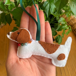May include: A white felt dachshund ornament with brown felt spots and a green ribbon. The ornament has a string of colorful beads around its neck.