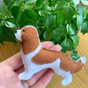May include: A handmade felt dog figurine in white and brown, with stitched details. The dog has brown ears and patches on its back, and a black nose. The figurine is held in a hand, with green foliage in the background.