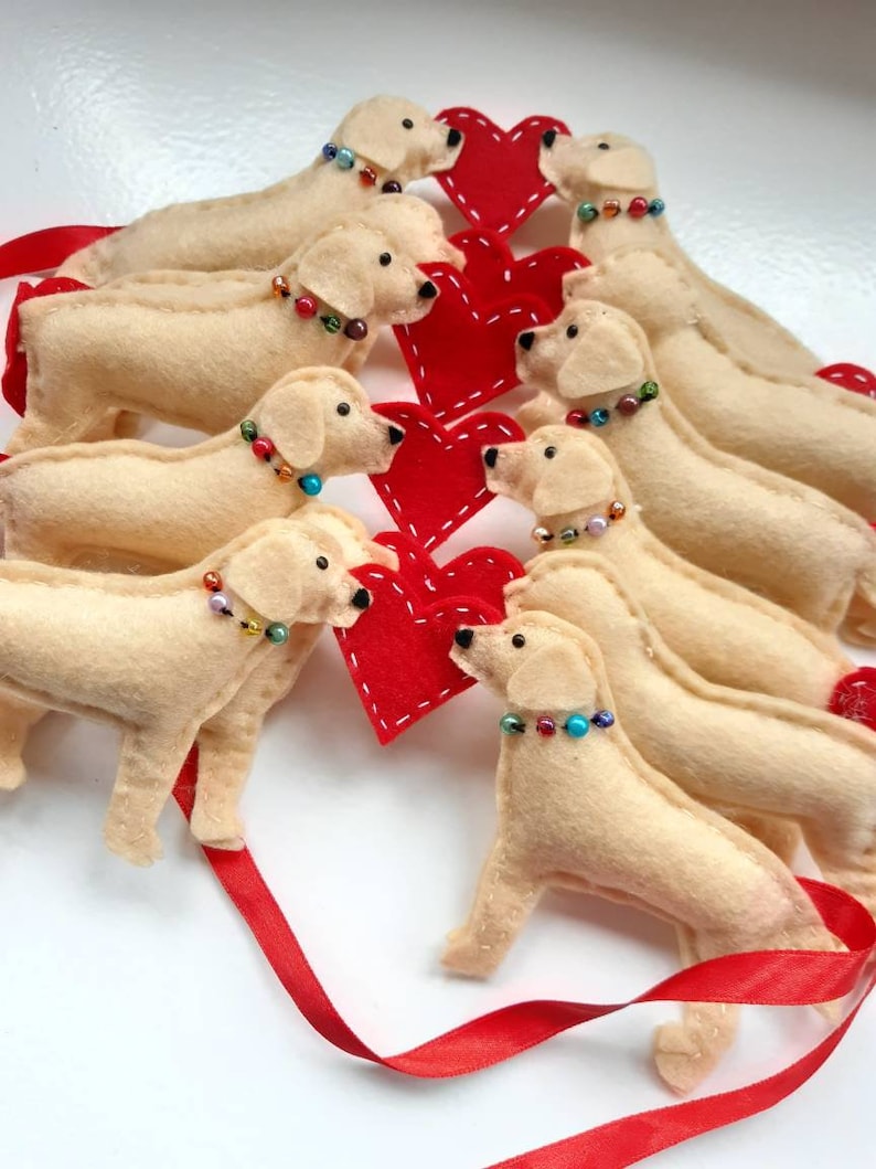 May include: A garland of felt golden retriever dogs with red felt hearts. The dogs are all facing the same direction and have beaded collars. The garland is tied with a red ribbon.