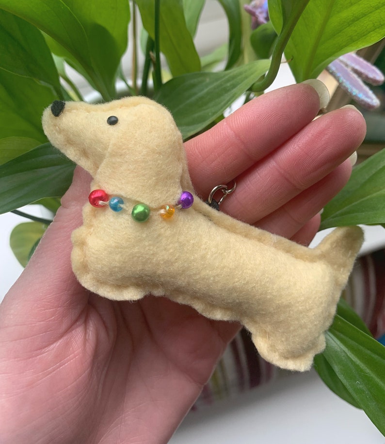 May include: A small, cream-colored felt dachshund keychain with a rainbow of beads around its neck.