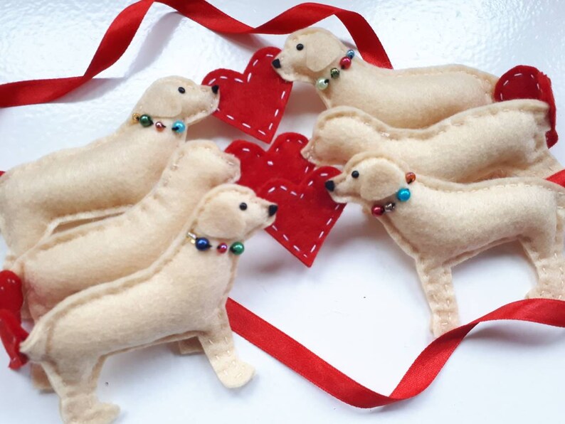 May include: Four felt golden retriever dog ornaments with red felt hearts. The dogs are arranged in a circle with their heads facing the center. Each dog has a beaded collar.