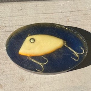 May include: A yellow and orange fishing lure encased in clear resin with a blue background. The lure has three hooks.