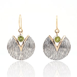 May include: A pair of silver and gold earrings with a green gemstone in the center of each earring. The earrings are shaped like a crescent moon with a V-shaped design in the center.