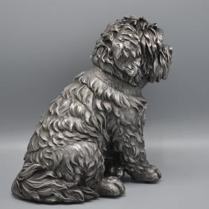 May include: A detailed silver-colored sculpture of a seated dog with a fluffy coat. The dog is depicted in a relaxed pose, with its head turned to the side. The sculpture has a collar and is set against a gray background.