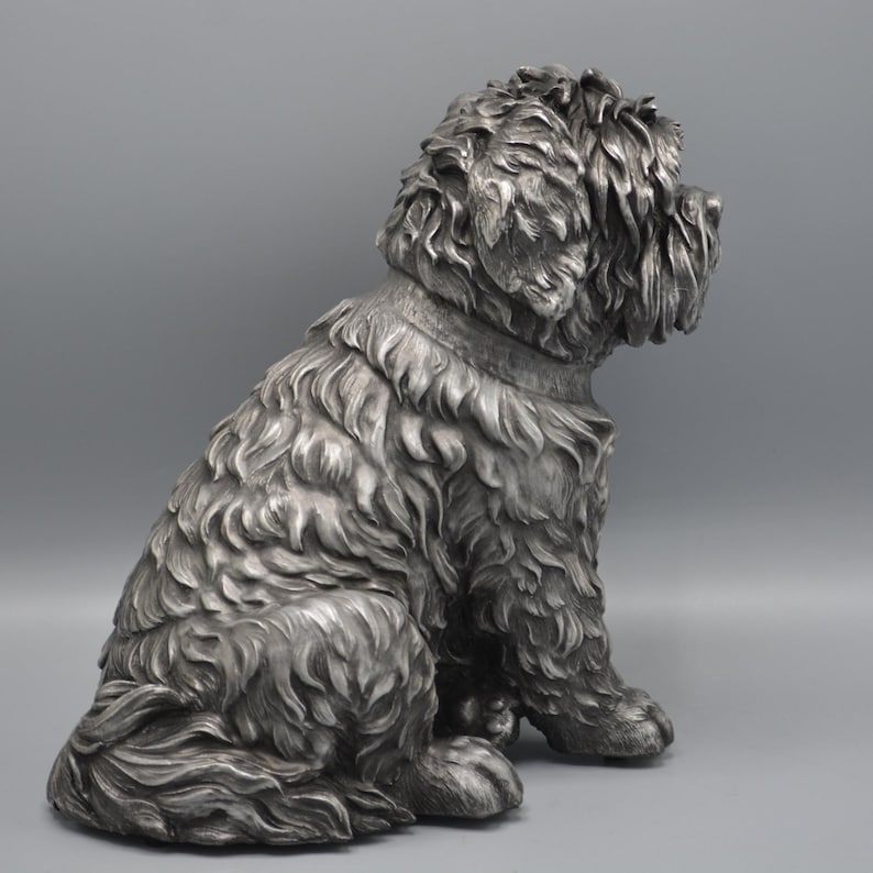 May include: A detailed silver-colored sculpture of a seated dog with a fluffy coat. The dog is depicted in a relaxed pose, with its head turned to the side. The sculpture has a collar and is set against a gray background.