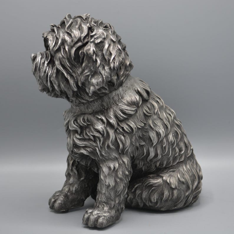 May include: A silver-colored, seated dog sculpture with a textured, curly coat. The dog is depicted in a relaxed pose, with its paws resting on the ground. The sculpture has a detailed, lifelike appearance, capturing the essence of a friendly canine companion.