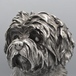 May include: A detailed, silver-colored sculpture of a dog's head. The dog has a fluffy, textured coat, with visible fur details. The sculpture captures the dog's facial features, including its eyes, nose, and mouth.