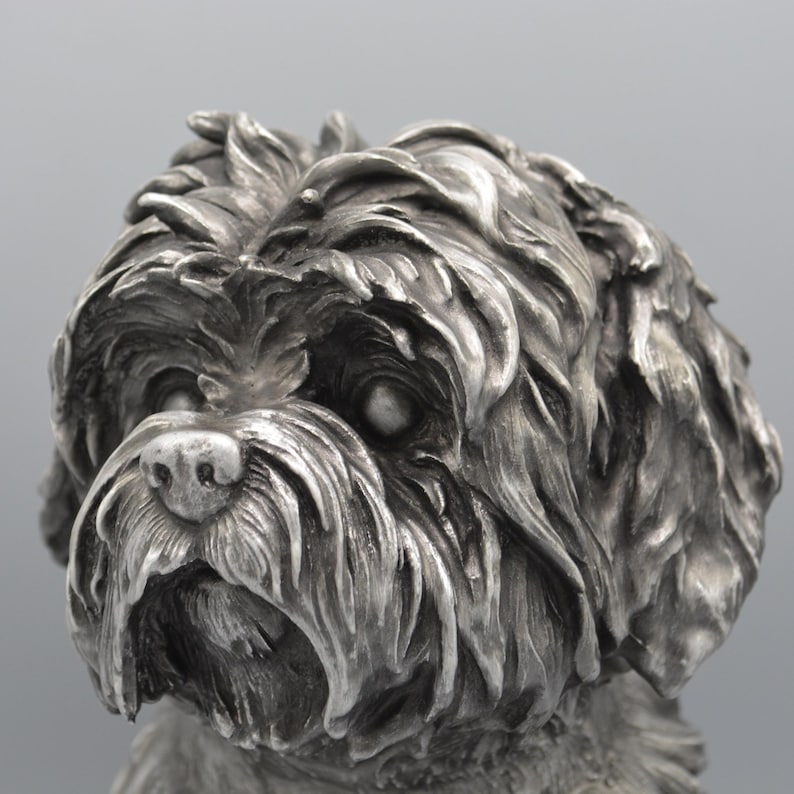 May include: A detailed, silver-colored sculpture of a dog's head. The dog has a fluffy, textured coat, with visible fur details. The sculpture captures the dog's facial features, including its eyes, nose, and mouth.