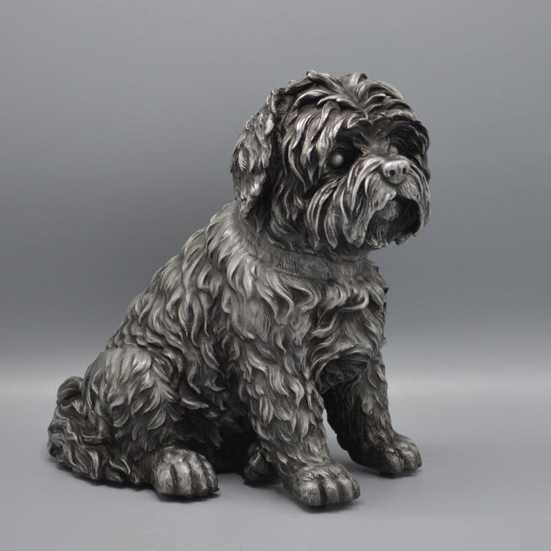 May include: A silver-colored dog statue in a seated position. The sculpture has detailed fur and a realistic depiction of a dog's face, with a focused gaze. The statue is on a solid gray background.
