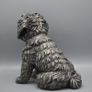 May include: A silver-colored dog statue in a seated position, viewed from the rear. The sculpture has detailed fur texture and a fluffy tail. The dog's head is turned to the left, with its ears slightly drooping.