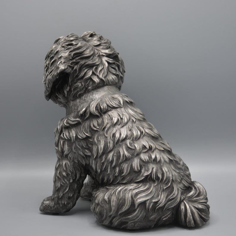 May include: A silver-colored dog statue in a seated position, viewed from the rear. The sculpture has detailed fur texture and a fluffy tail. The dog's head is turned to the left, with its ears slightly drooping.