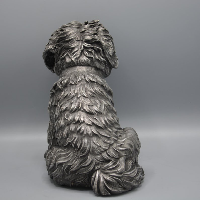 May include: A silver-colored dog statue, viewed from the back, sitting with its tail curled. The statue has detailed fur texture and a collar. The background is a solid gray.
