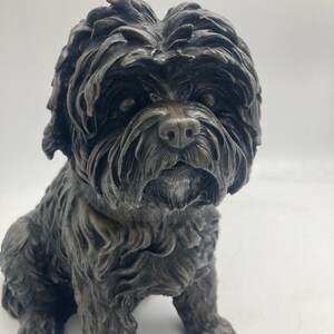 May include: A bronze-colored sculpture of a seated dog with a fluffy coat and detailed facial features. The sculpture has a textured surface and is labeled "Bronze" at the bottom.