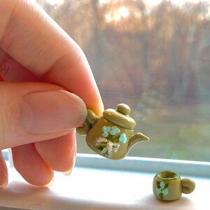 Puede incluir: Una tetera verde en miniatura con un diseño floral y una taza de té a juego. La tetera tiene un pico y un asa, y la taza de té tiene un asa. Ambas están pintadas con detalles verdes y azules.