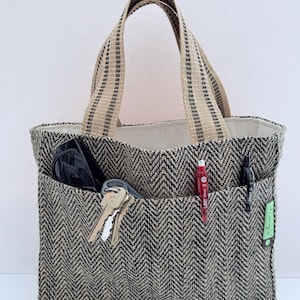 May include: A brown and black herringbone tote bag with two exterior pockets. The bag has a beige interior and beige and black striped handles. The pockets contain sunglasses, keys, and pens. A small green tag is attached to the side.