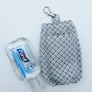 May include: A clear bottle of Purell Advanced Hand Sanitizer next to a gray and white patterned fabric hand sanitizer holder with a silver clip. The hand sanitizer bottle is partially visible, with the label facing the viewer.