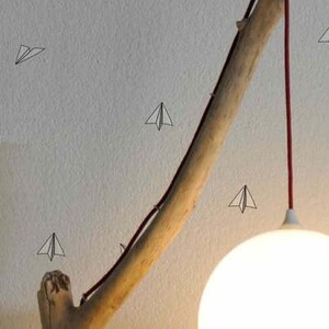 May include: A white globe lampshade hangs from a red cord attached to a branch-shaped floor lamp. The lamp is next to a large, off-white beanbag chair. The wall behind the lamp is decorated with small paper airplane stickers.