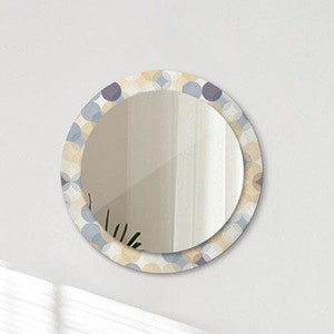 Op de afbeelding: Een ronde spiegel met een abstract patroonframe in wit, grijs en beige. De spiegel hangt aan een witte muur boven een bed met witte beddengoed en twee kussens.