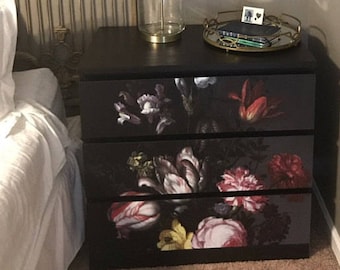 May include: A black dresser with three drawers features a floral print design. The drawers have a dark background with red, pink, and yellow flowers. A lamp with a white shade sits on top of the dresser.
