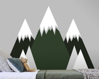 May include: A green and white mountain wall decal with the text "100% REMOVABLE BY StickersColoray" on a white circle sticker. The decal is on a gray wall behind a bed with a white and green blanket.