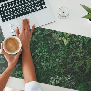 May include: A close-up of a person's hands holding a white coffee mug with a brown liquid inside. The mug is on a green and brown desk mat with a leafy pattern.