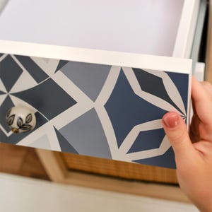 Può includere: Un cassetto bianco con un motivo geometrico nei toni del blu e del grigio. Il pomello è un piccolo pomolo rotondo con un disegno floreale. Il cassetto è parzialmente aperto, rivelando l'interno.