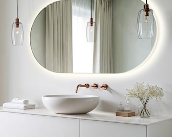 Puede incluir: Un tocador de baño ovalado blanco con un lavabo de cerámica blanca, un grifo de cobre y tres pomos de cobre. El tocador tiene dos cajones y está cubierto con una encimera blanca. Un espejo ovalado blanco con un borde iluminado cuelga sobre el lavabo. Dos lámparas colgantes de vidrio transparente con detalles de cobre cuelgan del techo. Un jarrón con flores blancas se encuentra sobre la encimera.