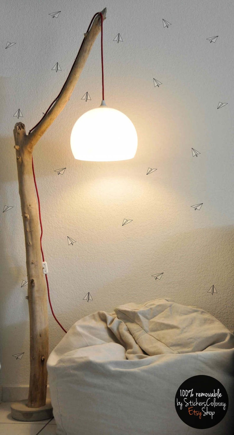 May include: A white globe lampshade hangs from a red cord attached to a branch-shaped floor lamp. The lamp is next to a large, off-white beanbag chair. The wall behind the lamp is decorated with small paper airplane stickers.
