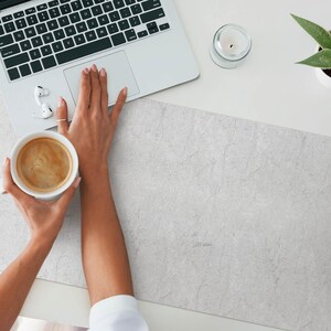 Op de afbeelding: Een witte keramische mok gevuld met koffie staat op een grijze, getextureerde bureauonderlegger. Een laptop, een kaars en een potplant met een succulent staan ook op het bureau.