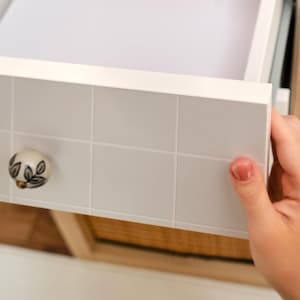 Peut inclure: Tiroir blanc avec un motif quadrillé et un bouton décoratif avec un motif floral. Le tiroir est partiellement ouvert, révélant l'intérieur. Le bouton a un fond crème avec des détails de feuilles vertes et noires.