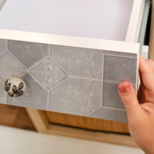 May include: A partially opened white drawer with a gray geometric tile design on the front. The drawer has a decorative knob with a floral design. The drawer is being opened by a person.