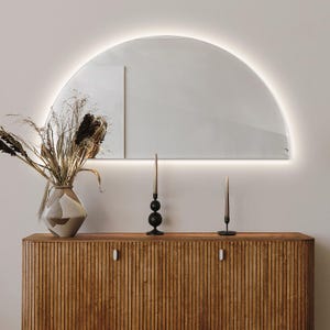 May include: A wooden cabinet with two doors and a half-circle mirror with a light strip around the edge. The mirror is mounted on a white wall above the cabinet. There are two black candle holders with black candles on the cabinet.