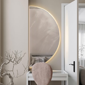 May include: A white vanity table with a white chair and a large, half-circle mirror with a light around the edge. The mirror is reflecting a bedroom with a bed and a window. There is a small, bare tree in the foreground.
