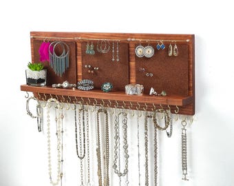 Könnte beinhalten: Ein wandmontierter Schmuck-Organizer aus Holz mit drei braunen Korkplatten für Ohrringe, einer horizontalen Metallstange, einer Ablage für Accessoires und einer Reihe goldfarbener Haken an der Unterseite für Halsketten und Armbänder.
