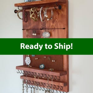 Peut inclure: Un organisateur de bijoux en bois avec une finition teintée foncée. Il comporte des étagères, des crochets et des tiges pour exposer colliers, bracelets et boucles d'oreilles. L'organisateur est fixé au mur et des bijoux y sont suspendus.