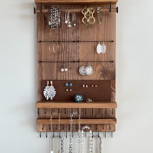 Puede incluir: Un organizador de joyas de madera con varios brazaletes, aretes y collares exhibidos. El organizador tiene un estante con una pequeña suculenta y objetos decorativos. La madera está teñida de un marrón cálido, y las joyas son plateadas y doradas.