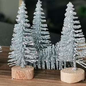 May include: A set of four miniature silver glitter Christmas trees with brown wooden bases. The trees are arranged in a row on a wooden surface.