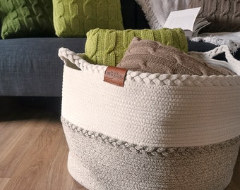 May include: A large, white and beige rope storage basket with a brown leather label that says "Cozy Vibes". The basket is filled with knitted blankets and pillows.