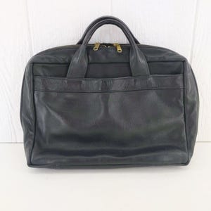 May include: Black leather briefcase with two top handles and a zippered closure. The bag has a front pocket and gold-colored zippers. The briefcase is rectangular and appears to be new.