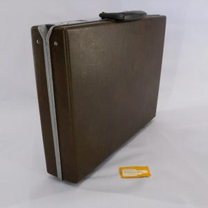 May include: A brown Samsonite briefcase with a silver trim and a black handle. The briefcase is rectangular and has a textured surface. A small Samsonite tag is on the ground.