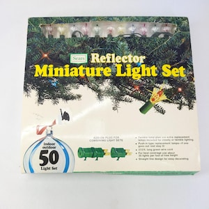 May include: A Sears Reflector Miniature Light Set box featuring a graphic of a Christmas tree with colourful lights. The packaging displays 50 indoor and outdoor lights, a 27.5 foot long green wire cord, and includes details about push-in replacement lamps.
