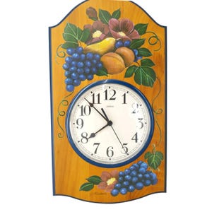 May include: A rectangular wall clock with a light wood finish and a blue border. The clock face features black Arabic numerals and the brand name Sunbeam. Hand-painted fruit motifs including purple grapes, pears, and peaches decorate the top and bottom.