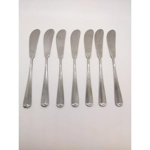 May include: A set of seven silver-toned stainless steel butter knives arranged in a row. Each knife features a wide, rounded blade and a slender handle that terminates in a subtle, decorative scalloped detail at the base of the handle.
