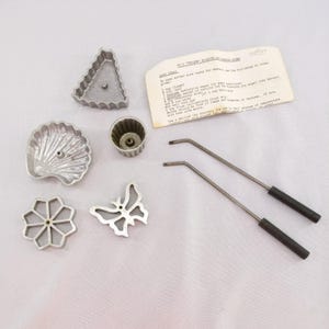May include: A collection of vintage aluminum waffle iron molds in various shapes, including a triangle, seashell, flower, butterfly, and a small cup. Two metal handles with black grips and a handwritten recipe card are also present.