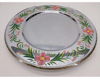 May include: A silver-colored decorative plate with a floral design. The rim features hand-painted pink flowers, green leaves, and small red accents. The plate has a gold-colored, scalloped edge.