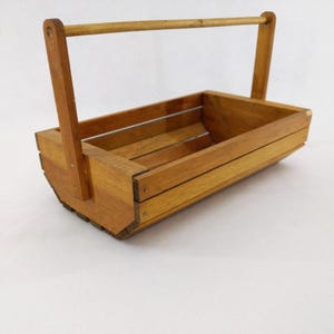 May include: A wooden basket with a rectangular body and a curved base. The basket has a handle that arches over the top. The wood has a warm, natural tone. The basket is likely used for carrying items.