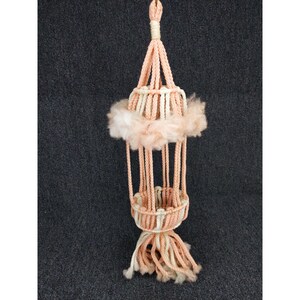 May include: A hanging macrame plant holder crafted from peach and cream-colored cord. The design features a central cylindrical structure with a fluffy, textured peach and cream fiber ring around the middle and a knotted tassel at the bottom.