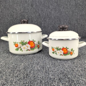 May include: Two cream-colored enamel pots with matching lids and side handles. Each pot features a decorative floral pattern with bright orange fruits, white blossoms, and green leaves. The lids have dark brown knobs and silver-toned rims.